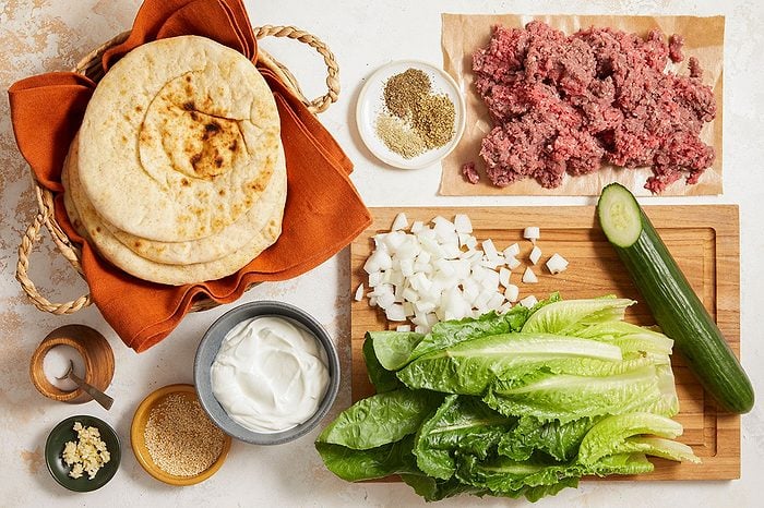 Overhead horizontal image of ingredients for Pita Burgers arranged on a light-colored surface. A woven basket holds several pita breads, next to a pile of ground beef on parchment paper. Chopped onions, whole lettuce leaves, a whole cucumber, minced garlic, a bowl of yogurt, sesame seeds, and various dried herbs and salt are arranged nearby on a cutting board and in small bowls.