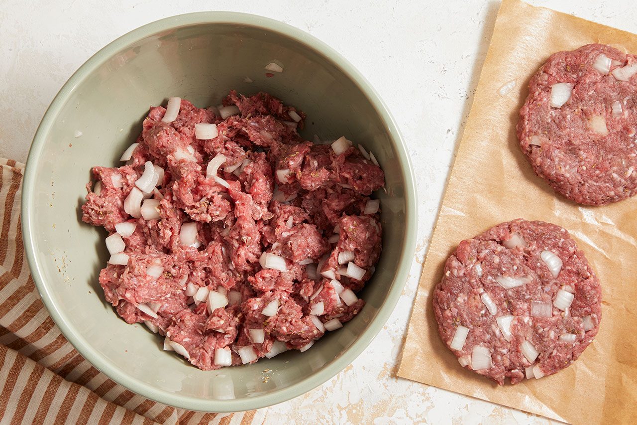 Overhead horizontal image of a pale green bowl containing a mixture of ground beef, chopped onions, and spices. To the right, two raw, formed burger patties, also made from the beef and onion mixture, rest on a piece of brown parchment paper on a white surface. A striped kitchen towel is visible in the bottom left corner.
