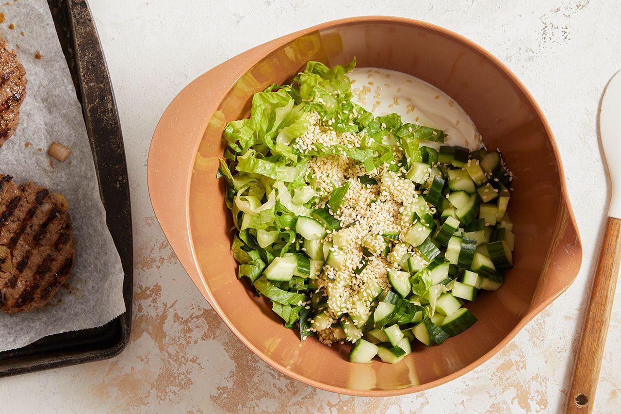 Overhead horizontal image of a brown mixing bowl filled with reduced-fat plain yogurt, shredded lettuce, chopped cucumber, and toasted sesame seeds. Partially visible on the left is a baking sheet with grilled lean ground beef patties.