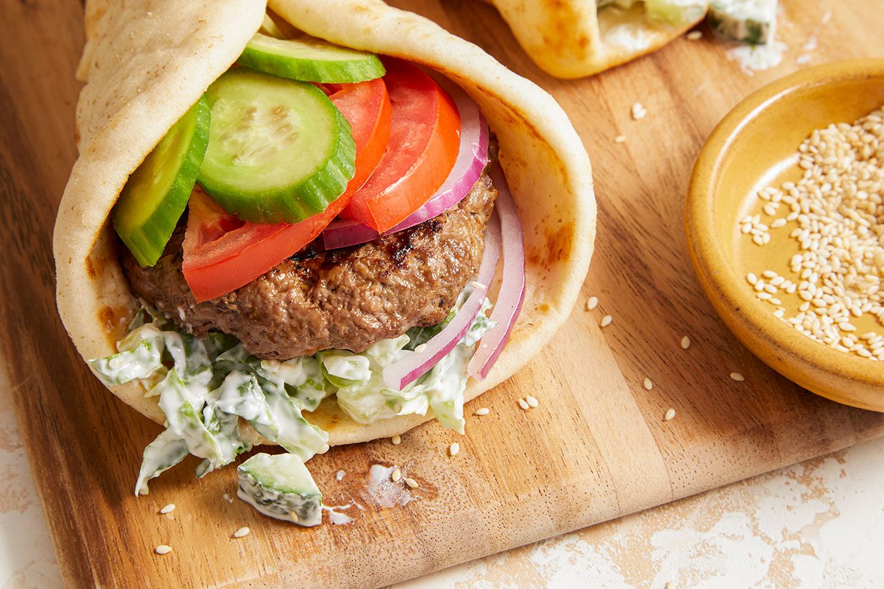 Eye Level horizontal image of an assembled Pita Burger placed on a wooden cutting board. The pita is filled with a lean ground beef patty, sliced cucumbers, sliced tomatoes, sliced red onion, and shredded lettuce with a creamy dressing. A small bowl of toasted sesame seeds is visible on the side.
