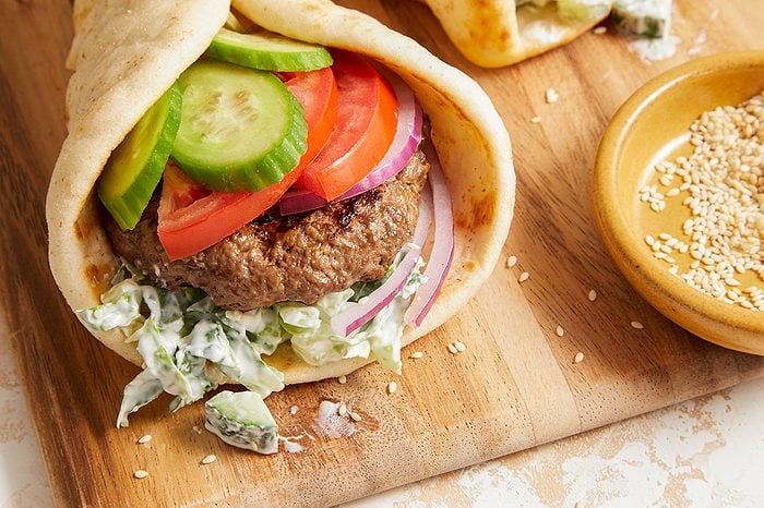Eye Level horizontal image of an assembled Pita Burger placed on a wooden cutting board. The pita is filled with a lean ground beef patty, sliced cucumbers, sliced tomatoes, sliced red onion, and shredded lettuce with a creamy dressing. A small bowl of toasted sesame seeds is visible on the side.