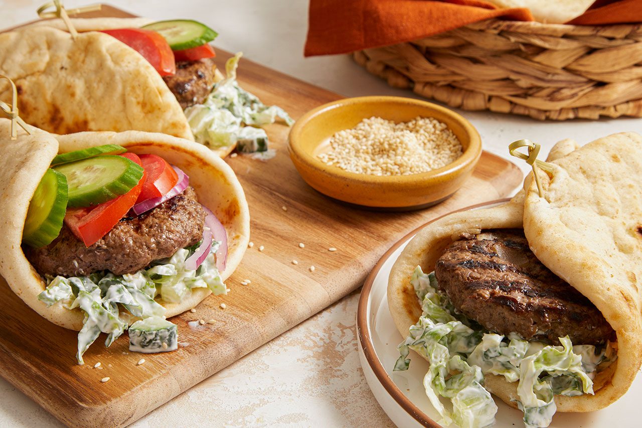 Eye Level horizontal image of two Pita Burgers presented on a wooden cutting board, with one burger on a plate. The burgers are assembled inside pita bread with a grilled beef patty, shredded lettuce with a creamy sauce, sliced cucumbers, sliced tomatoes, and sliced red onions. A small bowl of toasted sesame seeds is visible in the background, next to a wicker basket.