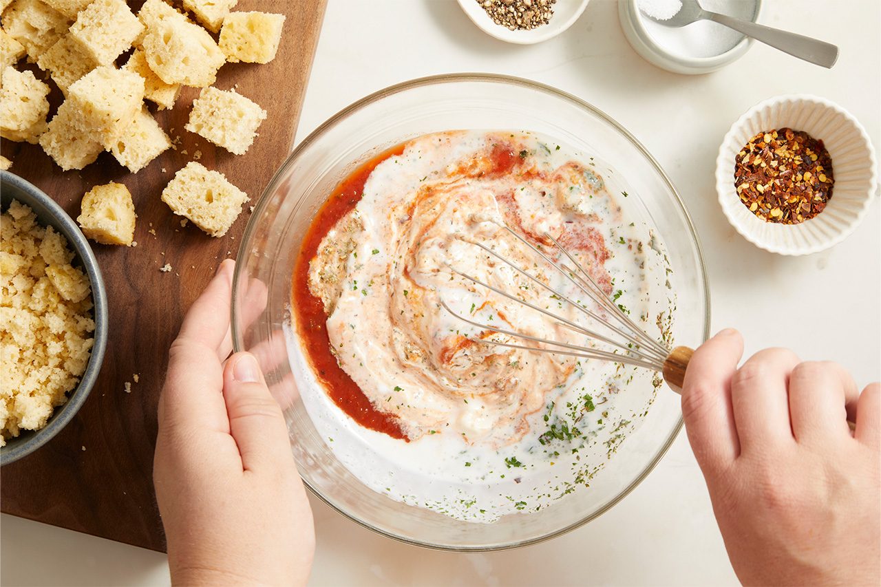 Overhead horizontal image of a person's hands whisking a creamy red sauce and herb mixture in a clear glass bowl. Nearby are cubed bread pieces on a wooden cutting board, crumbled bread crumbs in a small grey bowl, and small white dishes holding black pepper, salt, and red pepper flakes.