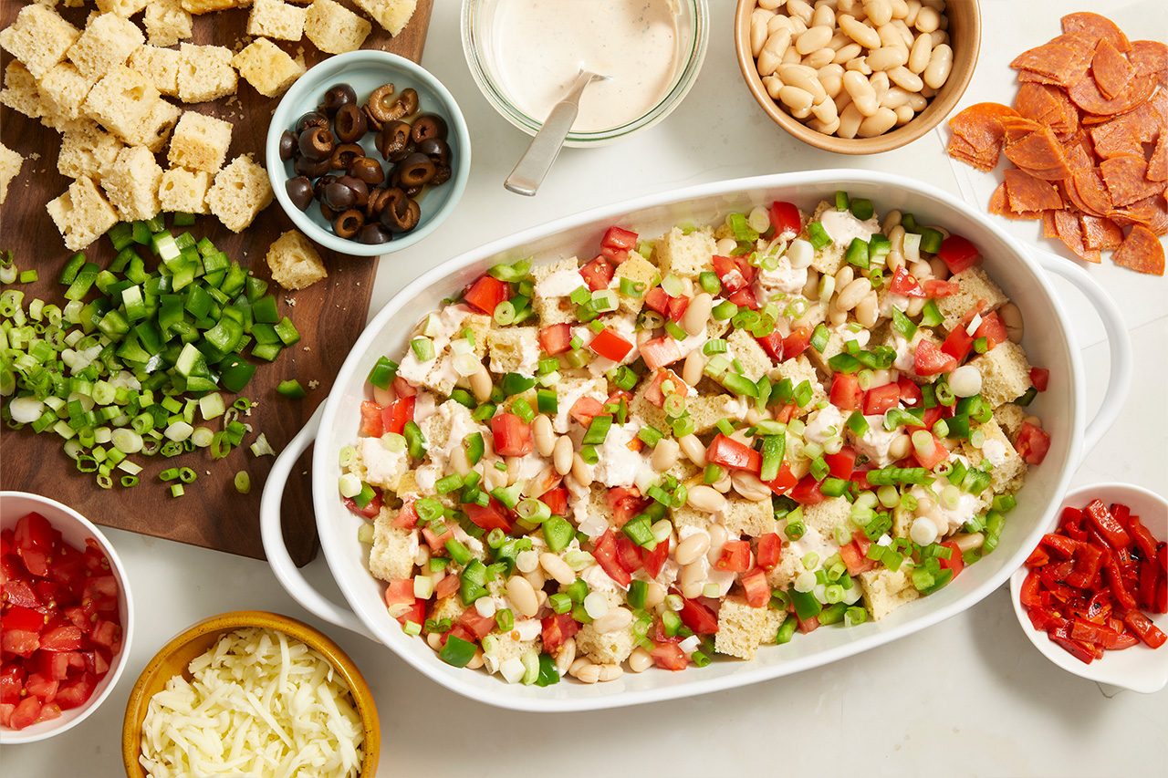 Overhead horizontal image of a white oval baking dish containing cubed bread, cannellini beans, diced tomatoes, diced green bell peppers, and chopped green onions, drizzled with dressing. Various prepped ingredients are arranged around the dish, including sliced black olives, more cubed bread, shredded cheese, sliced pepperoni, diced tomatoes, roasted red peppers, and a bowl of dressing.