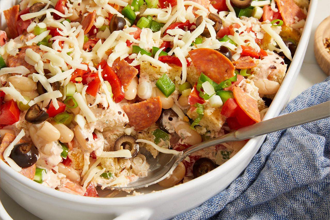 Overhead horizontal image of a casserole dish filled with a hearty mixture. The dish contains white beans, sliced pepperoni, chopped green bell peppers, diced tomatoes, sliced black olives, and bread pieces, all covered in a creamy sauce and topped with shredded mozzarella cheese. A metal spoon is partially submerged in the dish, ready for serving.