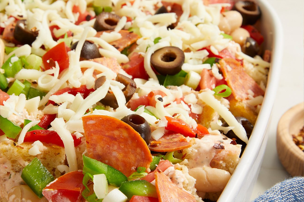 Eye Level horizontal image of an unbaked casserole in a white baking dish, richly topped with shredded mozzarella cheese, sliced black olives, diced tomatoes, diced green bell peppers, and sliced pepperoni. Chopped green onions and small white beans are also visible amidst the colorful ingredients.