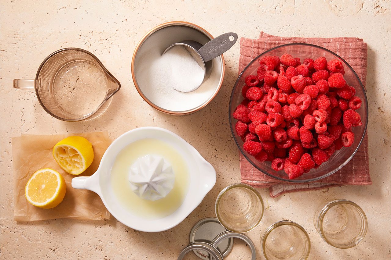 Overhead horizontal image of ingredients for canning, neatly arranged on a light-colored countertop. A large bowl of fresh raspberries, a cup of white granulated sugar, a measuring cup of clear liquid, and lemons with freshly squeezed lemon juice are visible, alongside empty canning jars and lids.