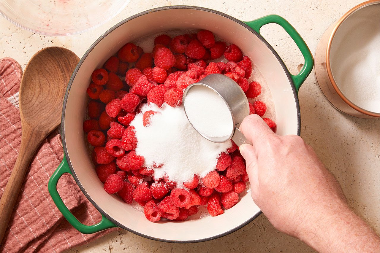 Overhead horizontal image of a hand adding granulated sugar from a metal measuring cup into a green enameled pot filled with fresh red raspberries. A wooden spoon rests on a pink kitchen towel beside the pot on a light countertop.