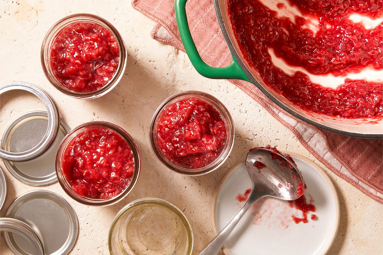 Overhead horizontal image of red fruit preserves being spooned into glass jars. Several jars are already filled with the chunky, berry-like jam, while others wait to be filled, alongside canning lids, a pot of remaining preserves, and a jam-covered ladle.