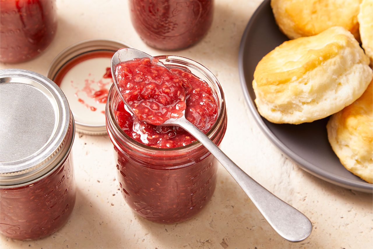 Eye level horizontal image of a spoon scooping raspberry jam from an open mason jar. More jars of jam are visible, along with a plate of golden biscuits to the right.
