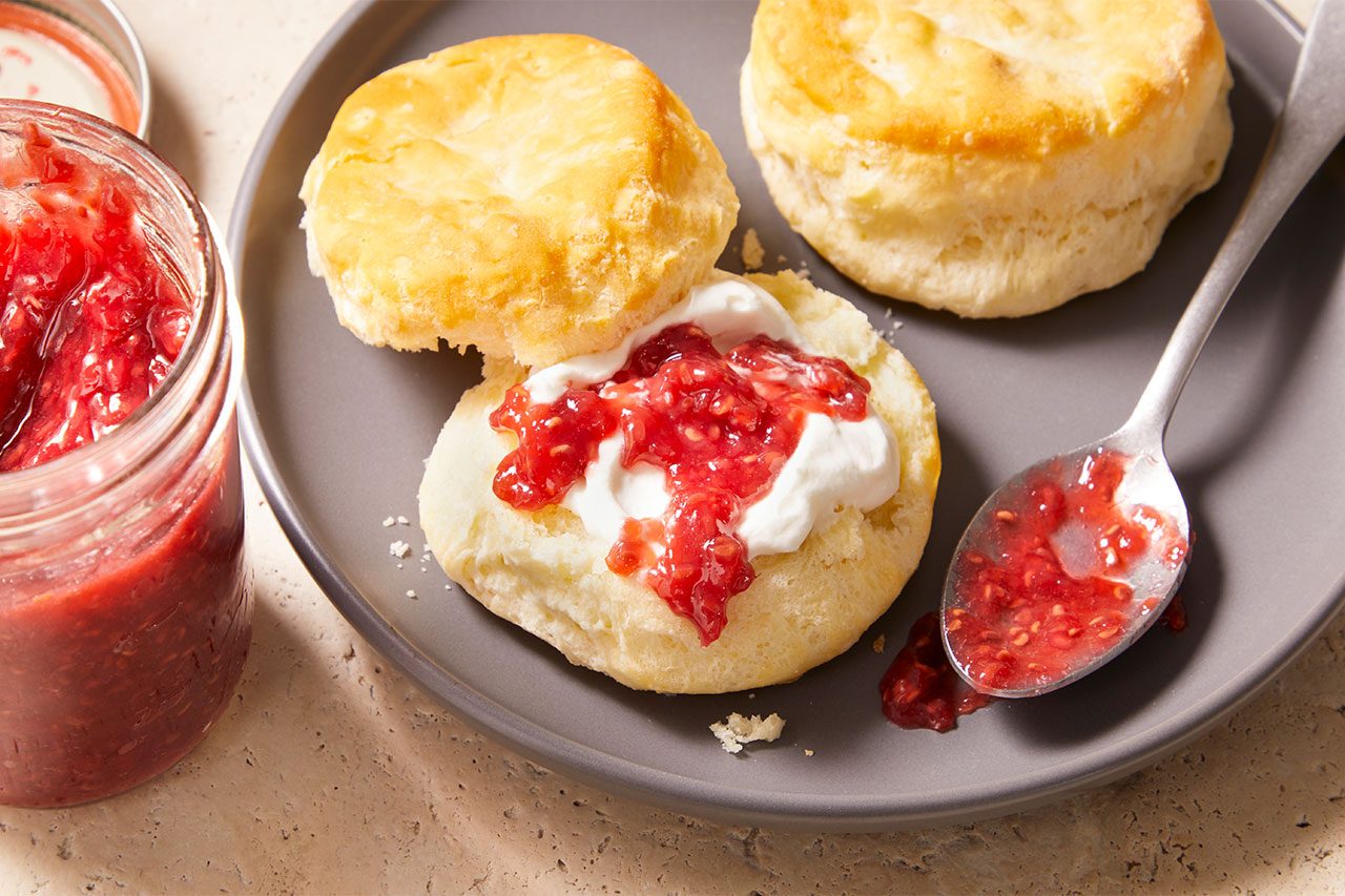 Overhead horizontal image of two golden-brown biscuits on a gray plate. One biscuit is split open and topped with a generous dollop of white cream and bright red raspberry jam, with a spoon of jam resting beside it and a jar of jam visible to the left.