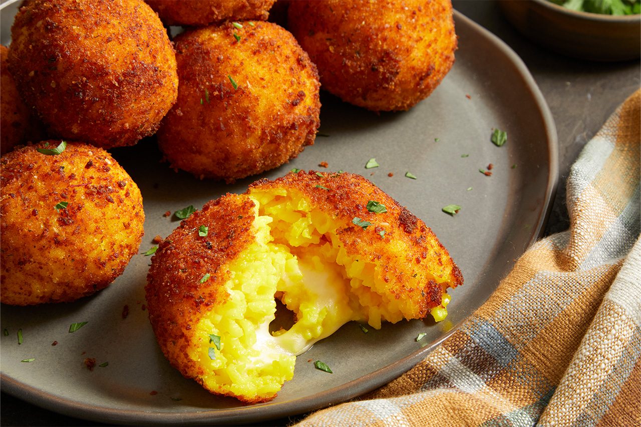Close-up shot of Saffron Arancini featuring a crispy golden-brown coating, with one arancini broken open to reveal vibrant saffron rice and a gooey melted cheese center, garnished with fresh parsley.