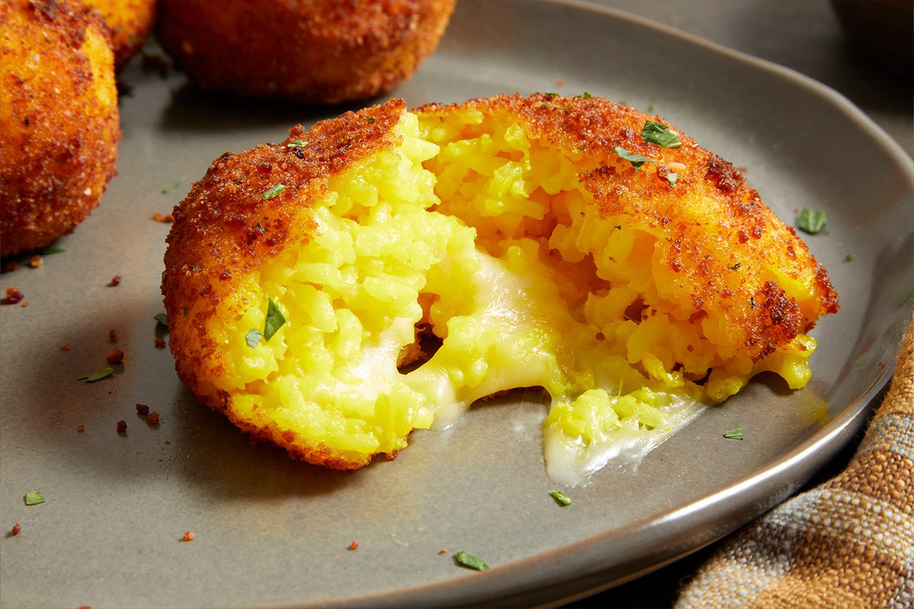 Close-up shot of Saffron Arancini broken open to reveal vibrant yellow saffron rice and a gooey melted cheese center, highlighting the crisp golden-brown coating and creamy filling, with additional arancini softly blurred in the background.