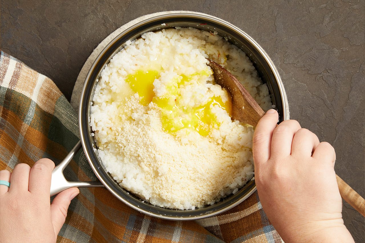 Overhead shot of cooked rice being mixed with grated cheese and melted butter in a saucepan using a wooden spoon, showing the preparation process for Saffron Arancini.