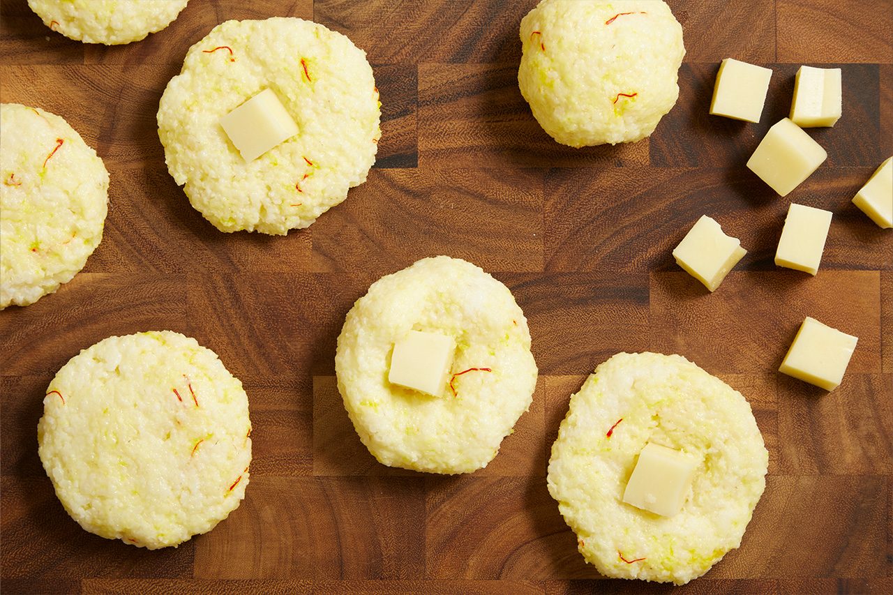 Overhead shot of saffron rice portions shaped into rounds on a wooden cutting board, each topped with a cube of cheese before forming the arancini, with additional cheese cubes scattered nearby.