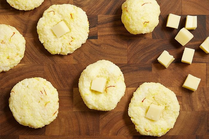 Overhead shot of saffron rice portions shaped into rounds on a wooden cutting board, each topped with a cube of cheese before forming the arancini, with additional cheese cubes scattered nearby.