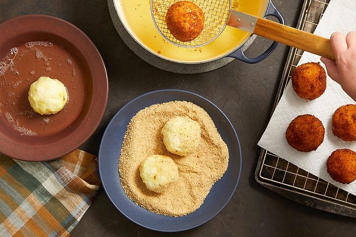 Overhead shot of Saffron Arancini being prepared and fried, featuring rice balls coated in breadcrumbs and one arancini being lifted from hot oil with a skimmer. Finished golden-brown arancini drain on a paper towel-lined tray nearby.
