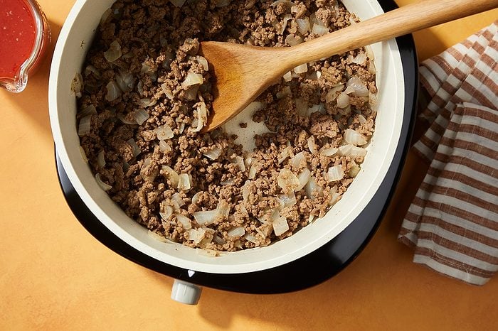 Ground beef and chopped onions being cooked in a white skillet, stirred with a wooden spoon. A striped kitchen towel and a small bowl of sauce sit nearby on an orange surface.