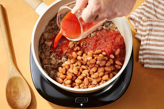 A hand pours tomato sauce into a pot containing beans and other ingredients on a stovetop, with a wooden spoon and striped towel nearby.