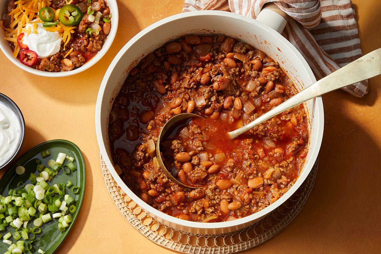 A pot of chili with ground meat and beans, a ladle inside, surrounded by bowls of toppings like shredded cheese, sliced jalapeños, sour cream, and chopped green onions on a tan surface.