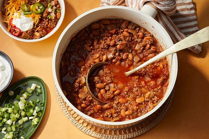 A pot of chili with ground meat and beans, a ladle inside, surrounded by bowls of toppings like shredded cheese, sliced jalapeños, sour cream, and chopped green onions on a tan surface.