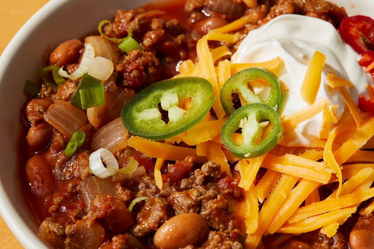 A bowl of chili topped with shredded cheddar cheese, sliced jalapeños, sour cream, green onions, and beans, with ground meat and tomatoes visible.