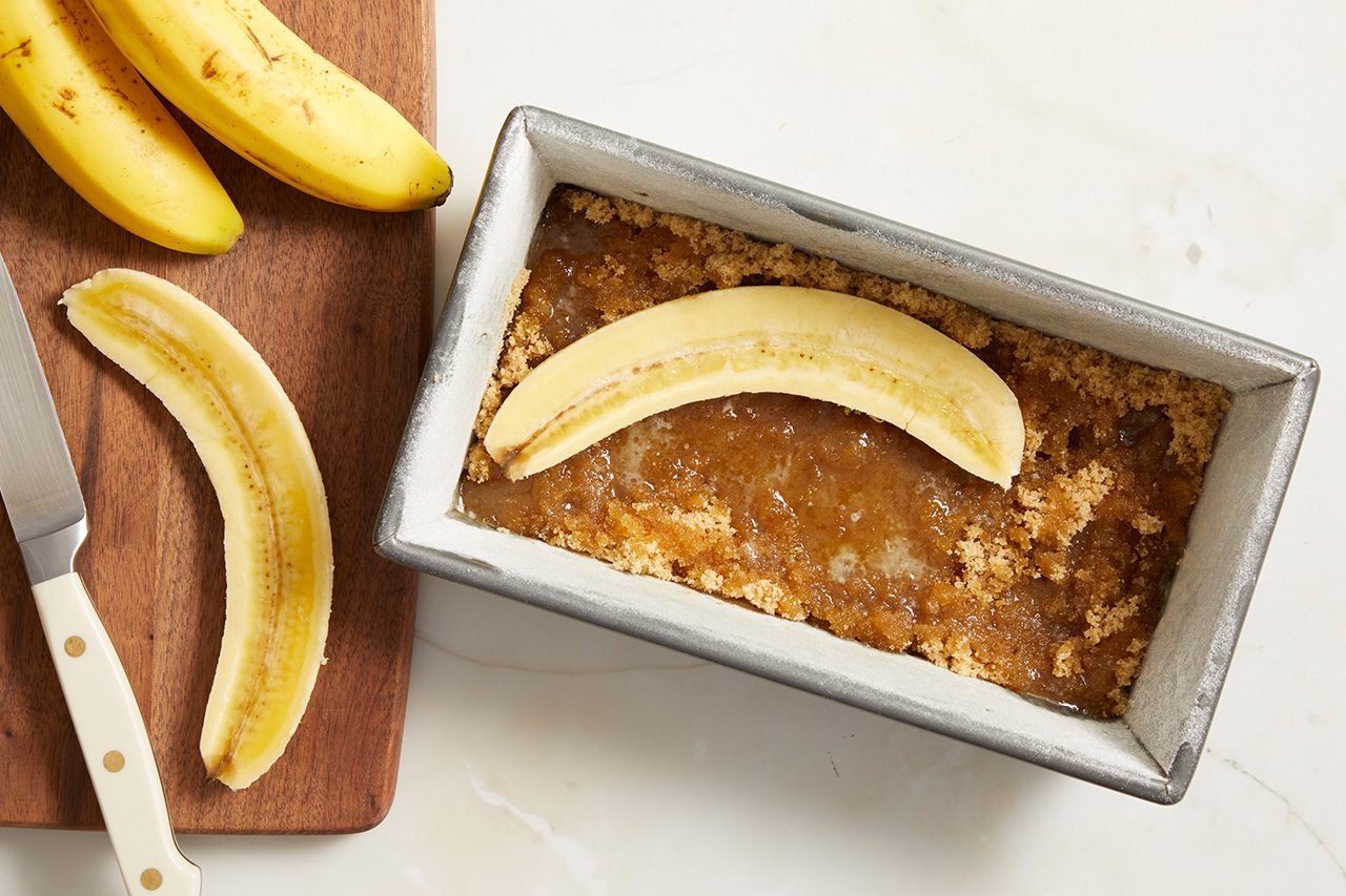 Overhead horizontal image of a peeled banana half placed on a brown sugar mixture in a metal loaf pan. On a wooden cutting board to the left, two whole bananas, another peeled banana half, and a knife are visible.