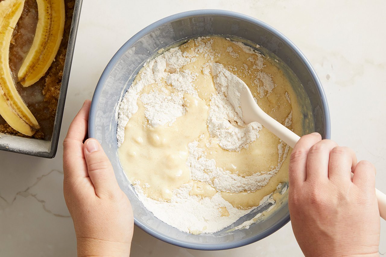 Overhead horizontal image of hands using a white spatula to mix dry flour into a wet batter in a blue mixing bowl. In the background, a loaf pan contains a banana-topped crumble mixture.