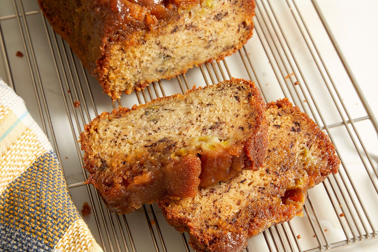 Overhead horizontal image of a freshly baked loaf of banana bread, partially sliced, cooling on a wire rack. The bread features a glossy, caramelized topping, with visible chunks of banana and dark specks throughout the tender crumb. A checkered tea towel is visible on the left.
