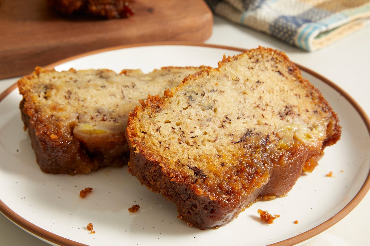 Eye Level horizontal image of two slices of freshly baked banana bread, featuring a caramelized topping and visible banana chunks, served on a white plate with a brown rim. The bread displays a moist texture and is speckled with dark bits, likely from ripe bananas.