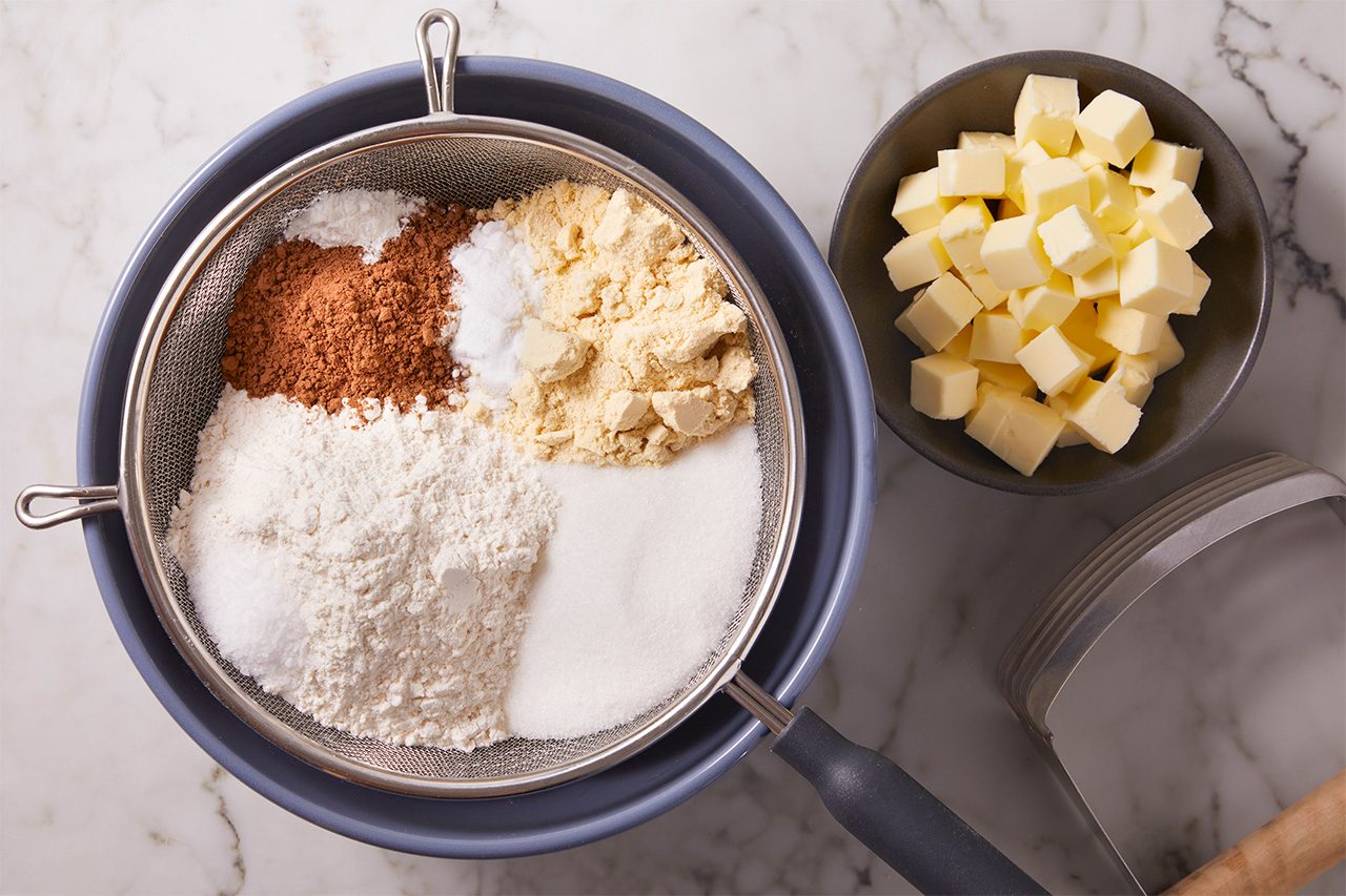 A sieve over a bowl holds flour, cocoa, sugar, baking powder, and salt. Next to it is a bowl of cubed butter, all on a marble countertop.