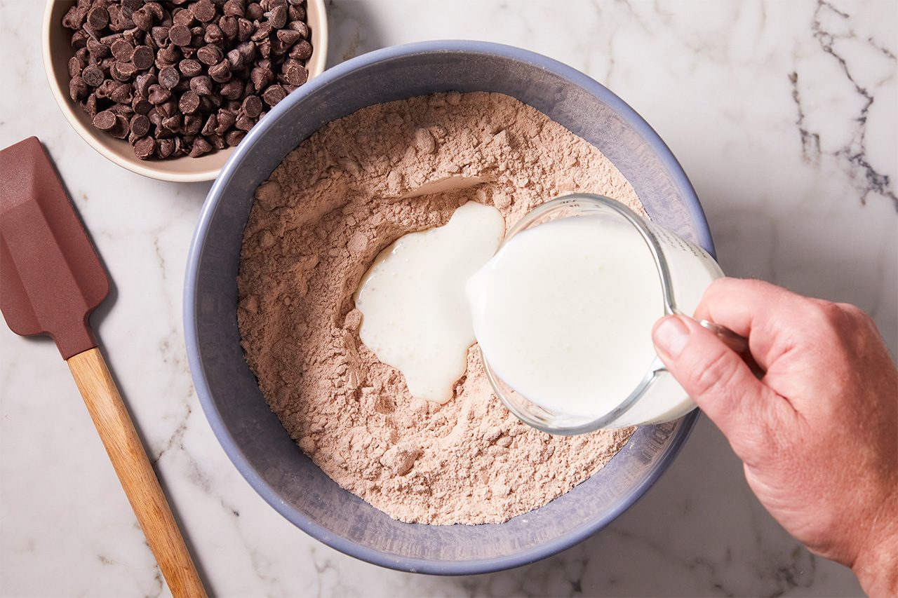 A hand pours milk into a bowl of chocolate cake mix on a marble counter, with a spatula and a bowl of chocolate chips nearby.