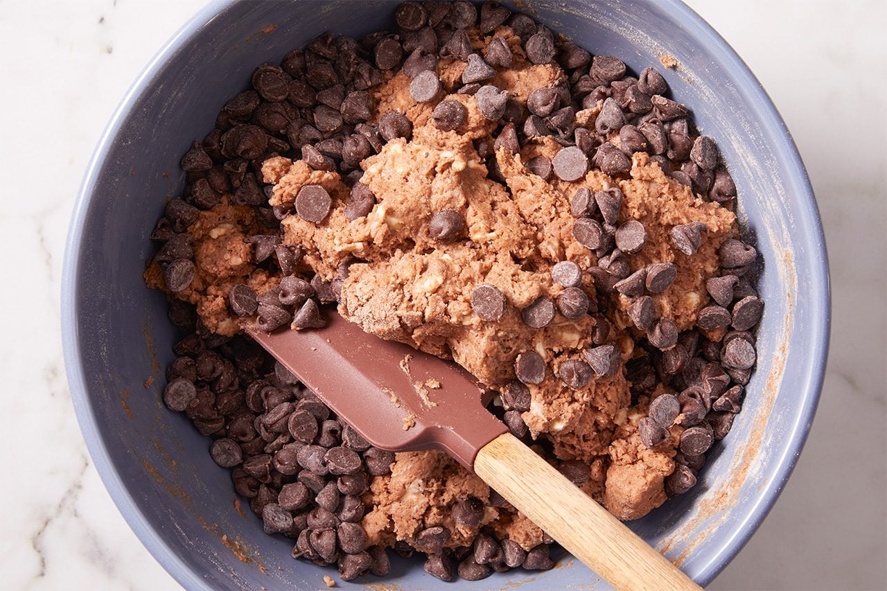 A mixing bowl with chocolate chip cookie dough and a spatula, showing chocolate chips mixed into the dough on a light surface.