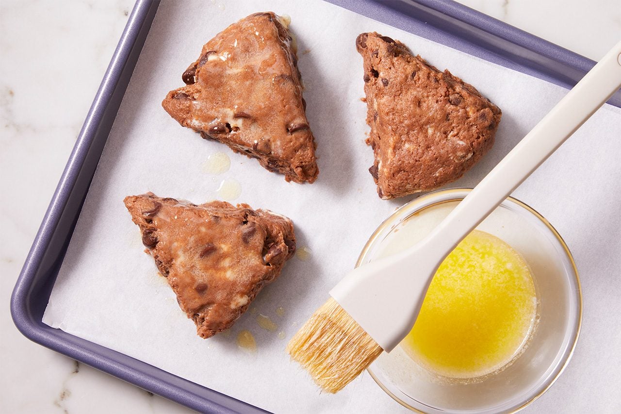 Three unbaked, triangular chocolate chip scones on a parchment-lined baking sheet, next to a bowl of melted butter with a pastry brush resting inside.