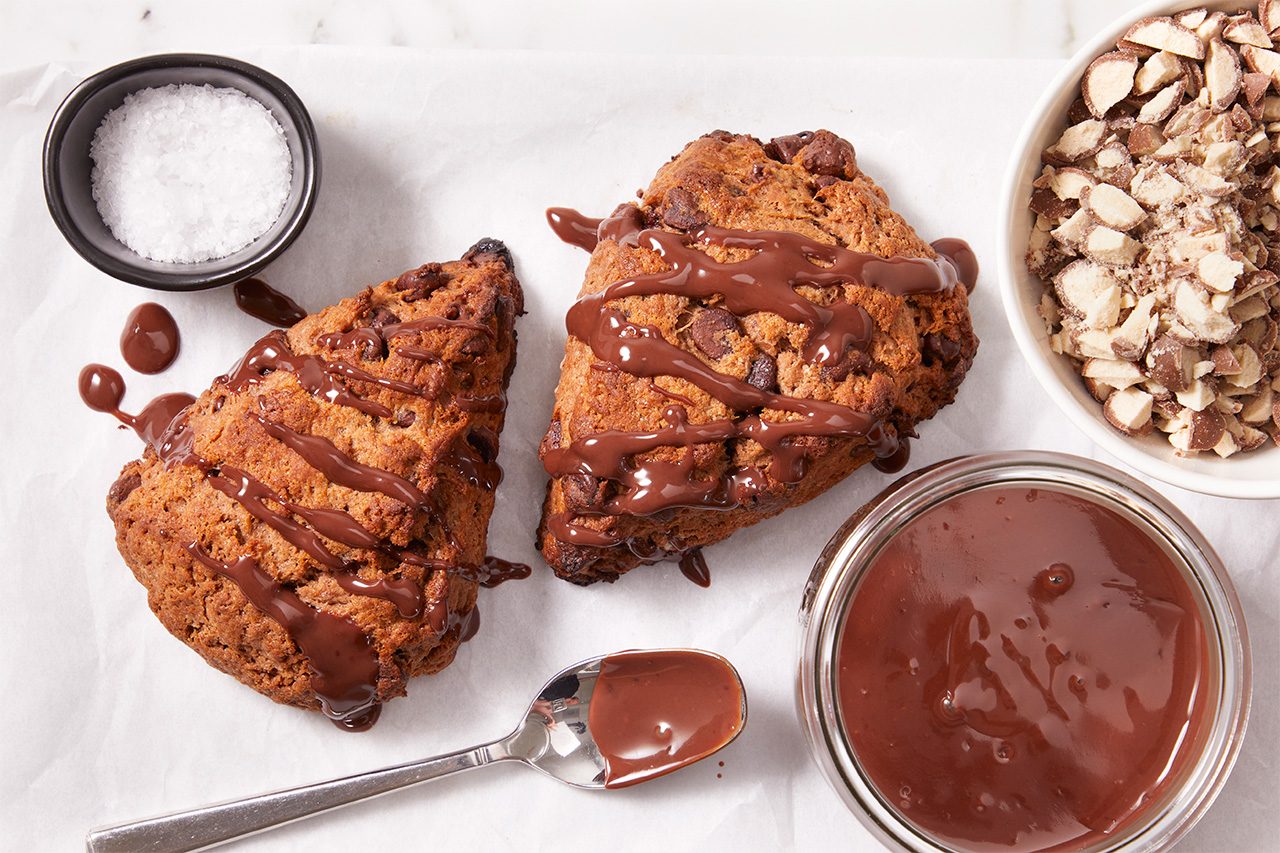 Two chocolate-drizzled scones on parchment, with a spoon, a bowl of melted chocolate, a bowl of chopped nuts, and a small dish of salt nearby.