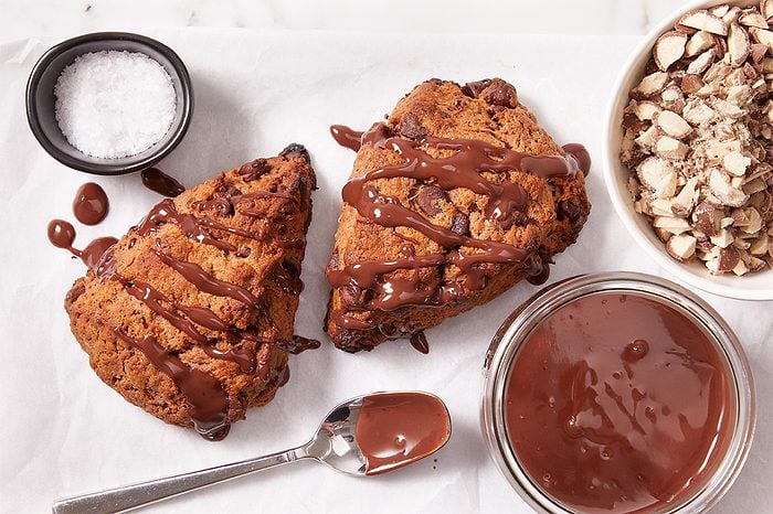 Two chocolate-drizzled scones on parchment, with a spoon, a bowl of melted chocolate, a bowl of chopped nuts, and a small dish of salt nearby.