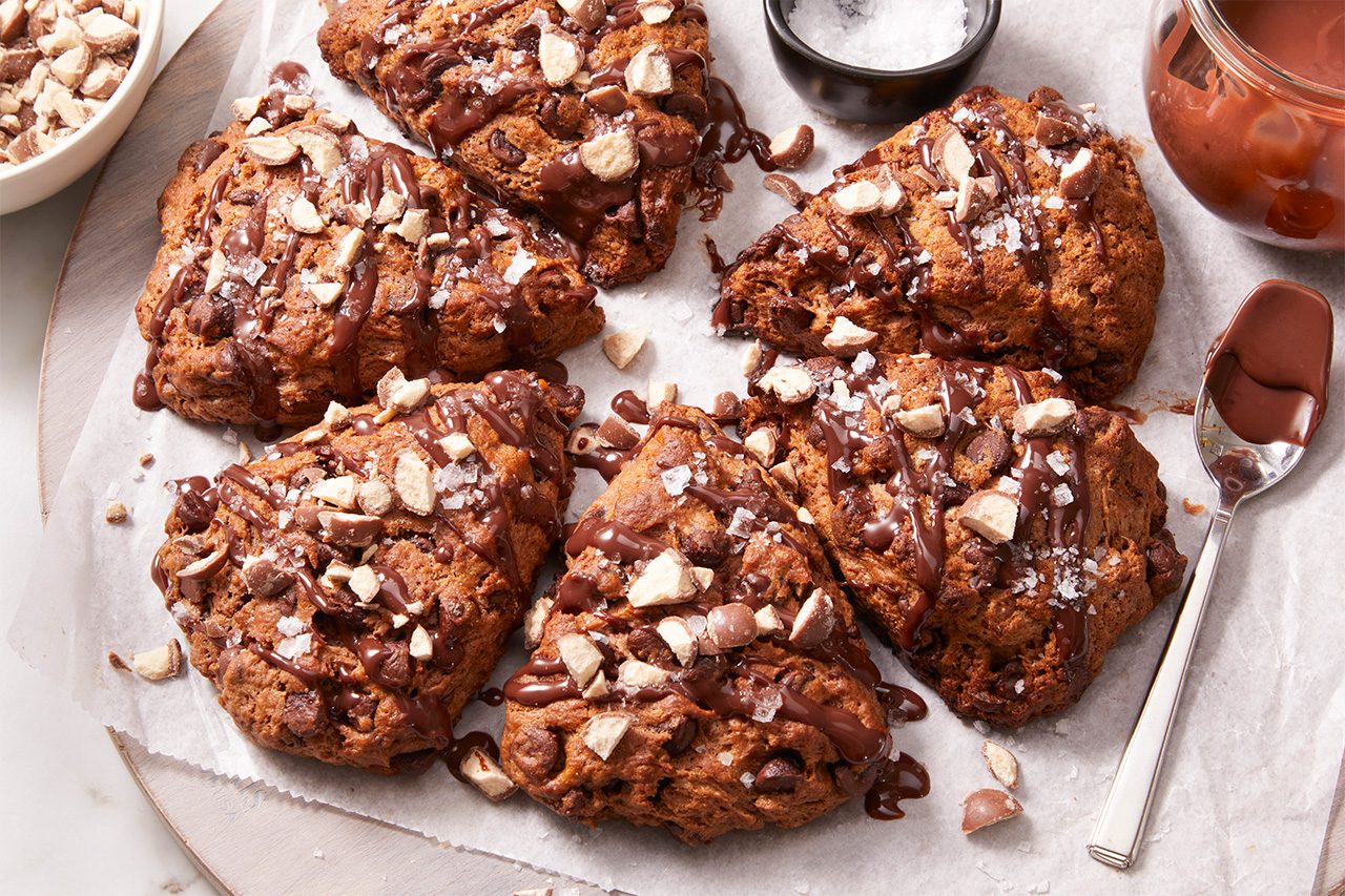 Six chocolate-drizzled scones topped with chopped nuts are arranged on parchment paper, with a spoon, a small bowl of sugar, and a jar of sauce nearby.