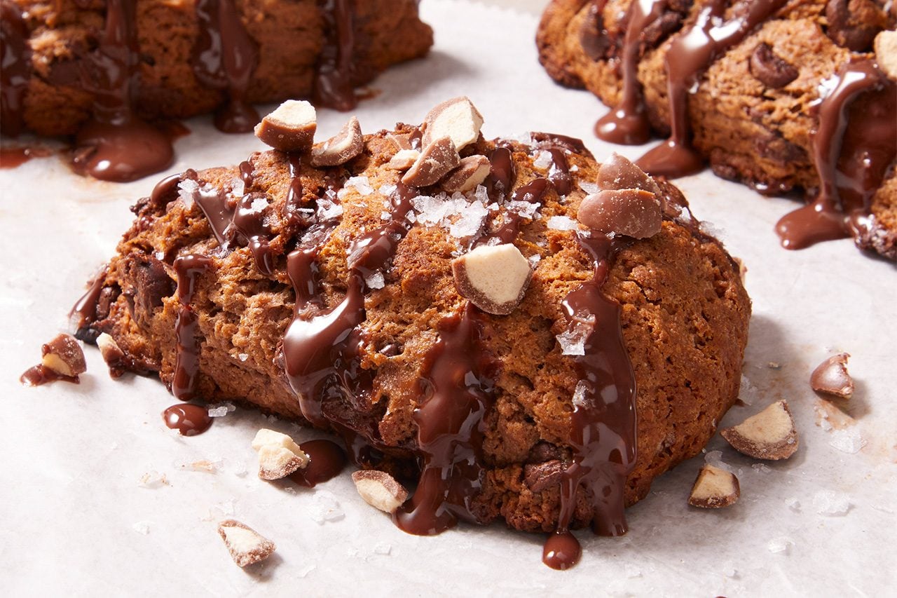 A chocolate chip scone drizzled with chocolate and topped with chopped nuts, resting on parchment paper.