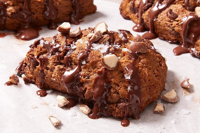 A chocolate chip scone drizzled with chocolate and topped with chopped nuts, resting on parchment paper.
