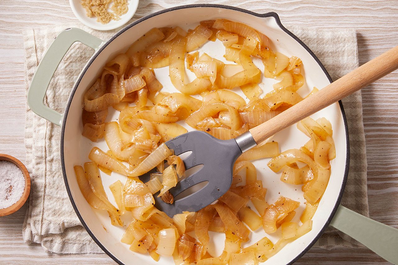 Overhead shot of caramelized onions cooking in a light-colored skillet — thinly sliced onions softened and turning golden-brown, coated in oil and spices, with a spatula gently stirring them on a rustic kitchen surface.