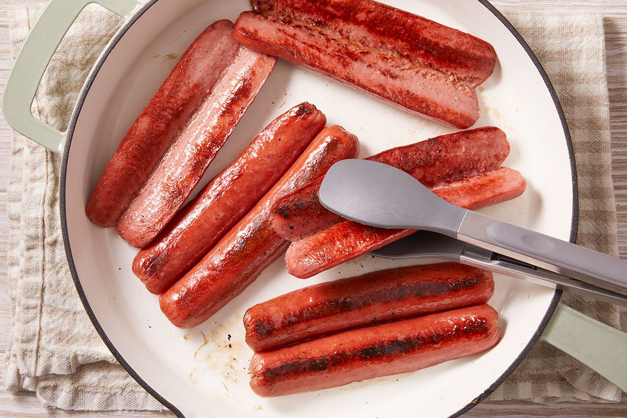 Close-up shot of sausages sizzling in a skillet; browned and slightly charred on the surface, glistening with oil, being turned with tongs as they cook evenly in a light-colored pan.