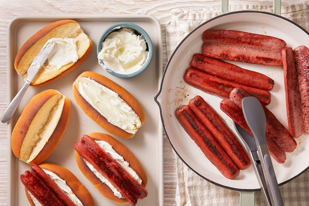 Overhead split composition of hot dog prep; toasted buns spread with a creamy layer on one side, while browned sausages rest in a skillet beside them. Some sausages are already placed into the buns, ready for assembly, with a clean, bright kitchen setup.