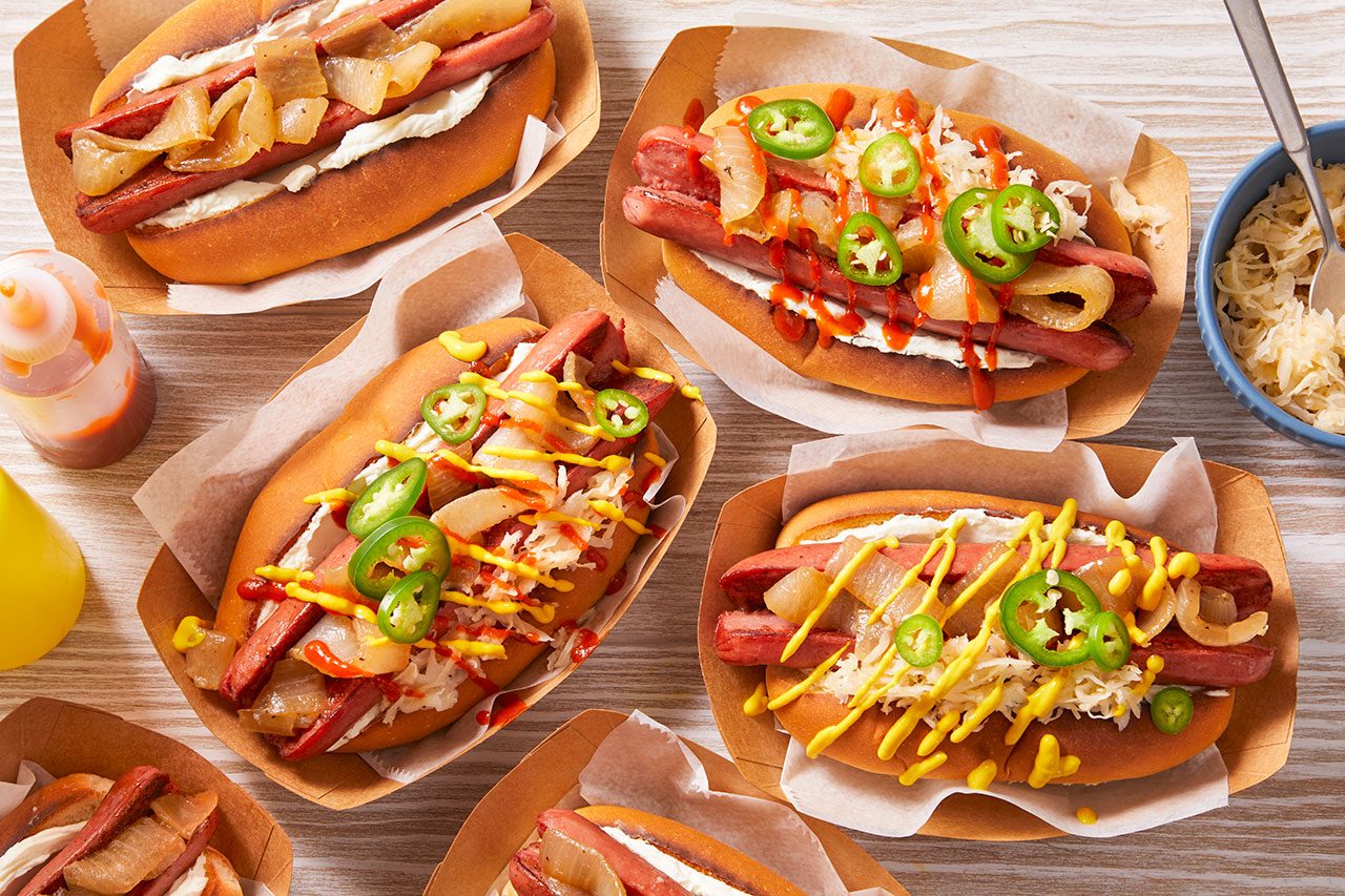 Overhead shot of assorted Seattle-style hot dogs arranged on a wooden surface, each in a toasted bun with grilled sausages, creamy spread, caramelized onions, melted cheese, and topped with jalapeño slices and drizzles of mustard and spicy sauce for a vibrant, loaded presentation.