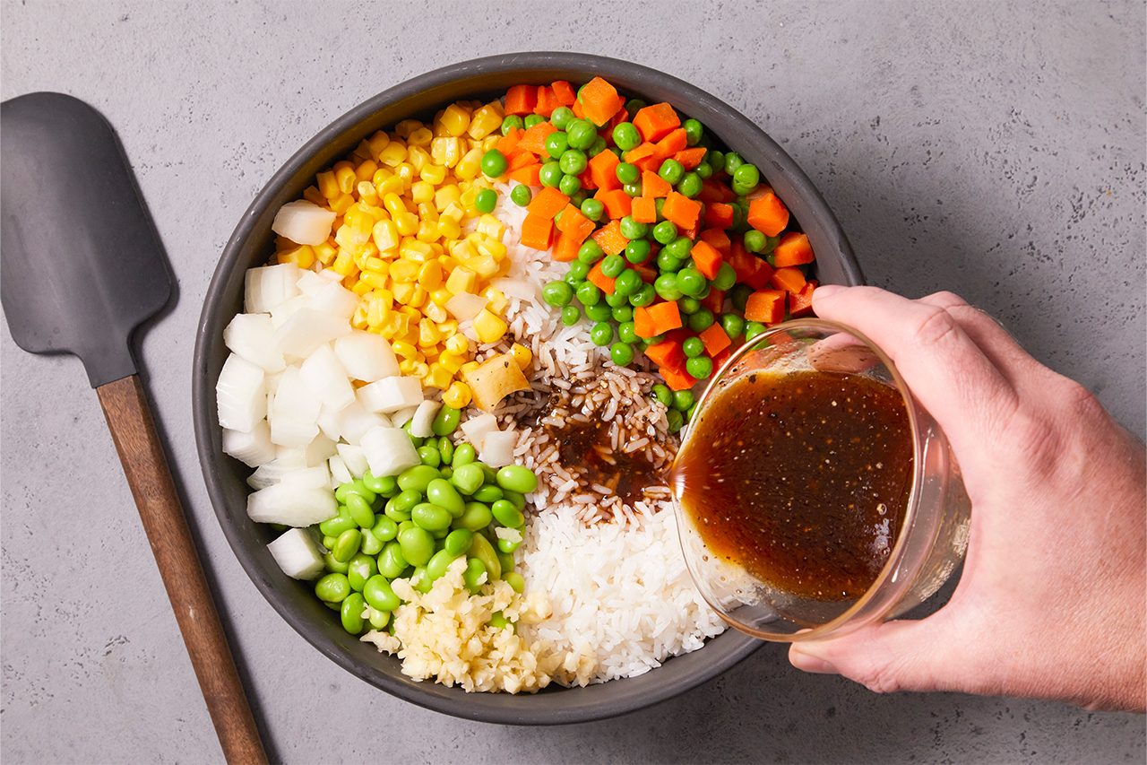 Overhead shot of a rice bowl being assembled; a base of white rice topped with neatly arranged vegetables like corn, peas, diced carrots, onions, edamame, and garlic, while a hand pours a savory sauce over the center for a fresh, colorful presentation.