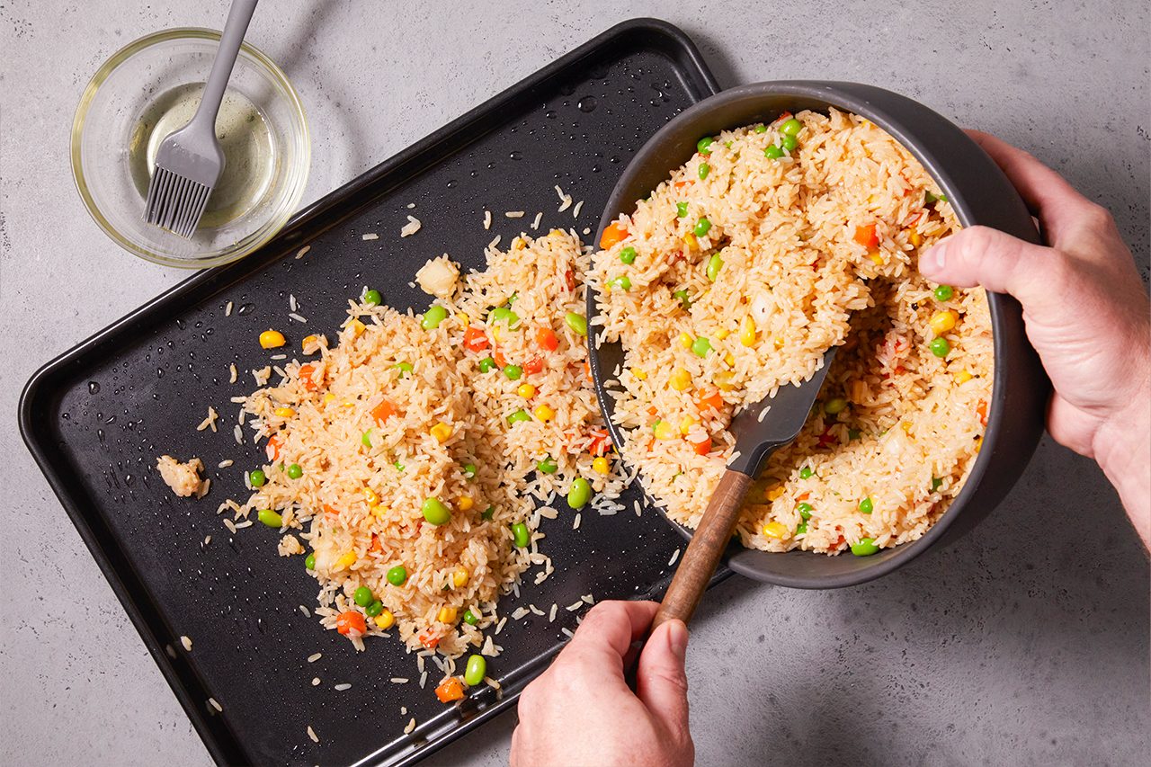 Overhead shot of fried rice being transferred from a bowl onto a baking tray, showing fluffy rice mixed with peas and carrots being spread out with a spatula, with a brush and oil placed nearby for preparation.