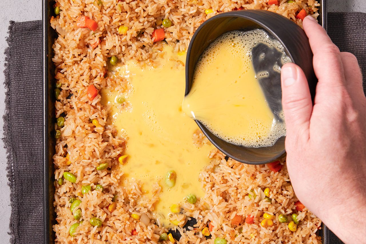 Overhead shot of fried rice spread across a baking tray as a hand pours beaten eggs into the center, creating a rich layer that begins to blend with the rice and mixed vegetables like peas, carrots, and corn for a hearty, oven-style dish.