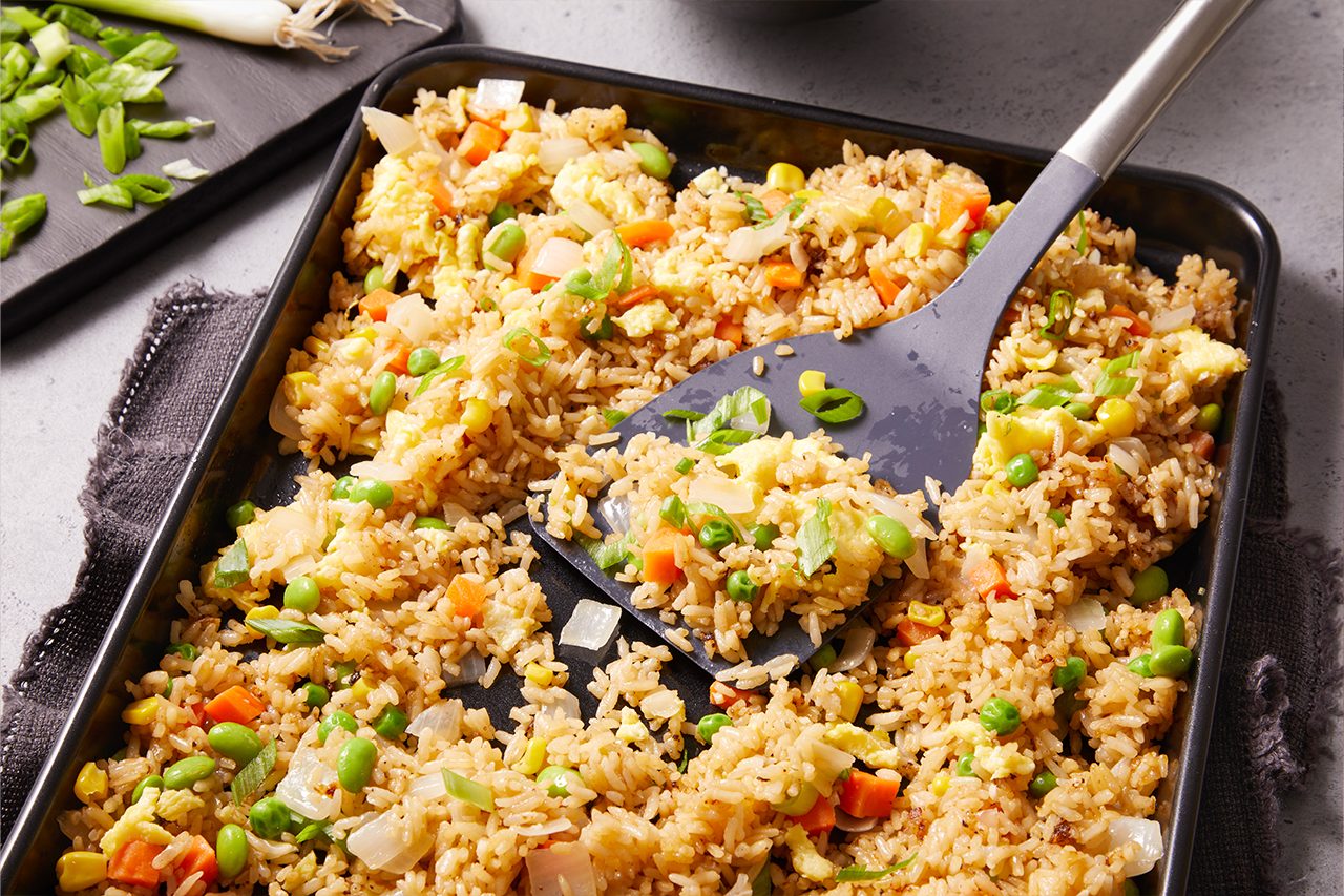 3/4 angle view shot of sheet-pan fried rice, showing golden rice mixed with peas, carrots, and onions, with a spatula lifting a portion from the tray, highlighting its fluffy texture and evenly cooked ingredients.