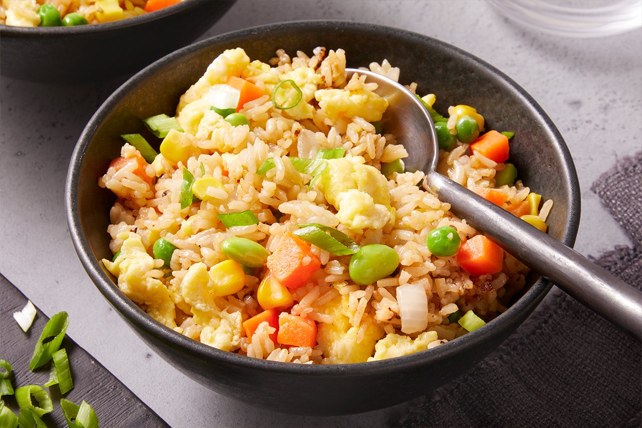 Close-up shot of a bowl of sheet-pan fried rice, featuring fluffy grains mixed with scrambled eggs, peas, carrots, corn, and edamame, topped with fresh green onions and served with a spoon for a warm, comforting presentation.