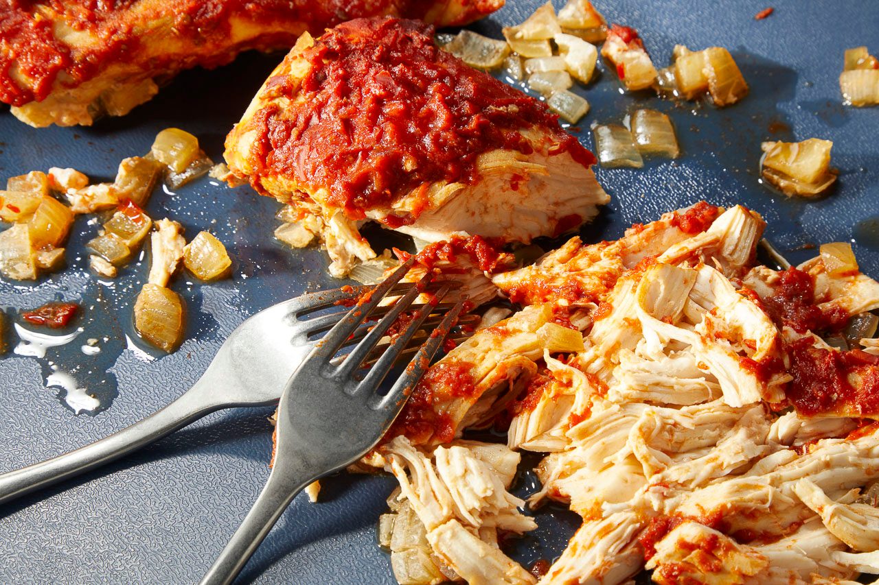 Overhead horizontal image of chicken being shredded and mixed with red sauce and sautéed onions using a spoon and a fork