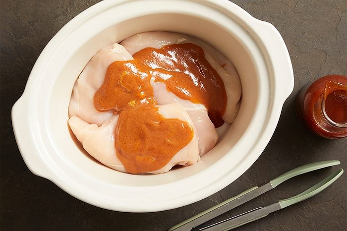 Raw chicken pieces in a white slow cooker with reddish-brown sauce on top; metal tongs and a small jar of sauce sit nearby on a dark countertop.